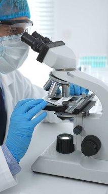 Koruyucu maske takan kıdemli erkek bilim adamı laboratuarda mikroskop kullanarak tıbbi araştırma ve analiz yapıyor. Tıp, sağlık ve bilim konsepti.