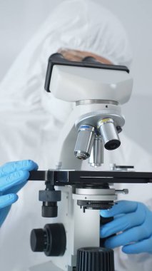 Tehlikeli madde giysisi, maske ve mavi eldiven giyen erkek bir virüs bilimci laboratuvarda virüsleri araştırırken mikroskop altında özenle çalışıyor. Tıp ve bilim konsepti.