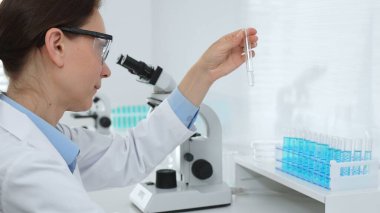 Laboratuvar önlüğü giyen ve koruyucu gözlük takan kadın bilim adamı modern laboratuarda mikroskop ve test tüpleri askısı ile test tüpünü temiz sıvıyla analiz ediyor. Tıp, sağlık ve bilim konsepti