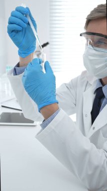 Laboratuvar önlüklü, koruyucu gözlüklü ve mavi eldivenli maskeli erkek bilim adamı modern laboratuardaki test tüplerine mikroskop ve test tüpleri rafı koyuyor. Tıp, sağlık ve bilim konsepti.