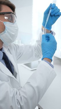 Laboratuvar önlüklü, koruyucu gözlüklü ve mavi eldivenli maskeli erkek bilim adamı modern laboratuardaki test tüplerine mikroskop ve test tüpleri rafı koyuyor. Tıp, sağlık ve bilim konsepti.