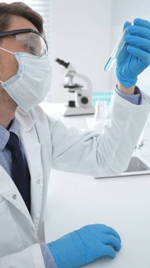Koruyucu giysi giyen bilim araştırmacısı modern laboratuvar çalışma alanında mavi sıvı numunesini test tüpünden inceliyor. Tıp, sağlık ve bilim konsepti