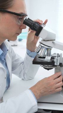 Laboratuvar önlüğü ve koruyucu gözlüklü kadın bilim adamı modern bir laboratuvarda mikroskop altında test örnekleri inceliyor. Tıp, sağlık ve bilim konsepti