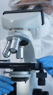 Tehlikeli madde giysisi, maskesi ve eldiveni olan bir tıp virologu bir laboratuvar araştırma virüsünün mikroskobunda özenle çalışıyor. Sağlık ve Bilim Konsepti.