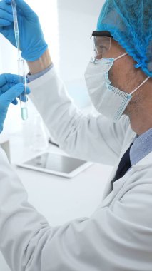 Laboratuvar şapkası, yüz maskesi, Google ve mavi eldiven takan erkek tıp bilimci modern bir laboratuvar ortamında bir deney tüpünün içine dikkatlice sıvı bırakıyor. Tıp, sağlık hizmeti kavramı.
