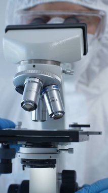 Tehlikeli madde giysisi, maskesi ve eldivenleri olan bir tıp virologu laboratuvar araştırmalarında mikroskop başında özenle çalışıyor. Tıp ve bilim konsepti.