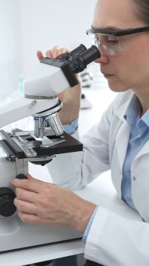 Laboratuvar önlüğü ve koruyucu gözlüklü kadın bilim adamı modern bir laboratuvarda mikroskop altında test örnekleri inceliyor. Tıp, sağlık ve bilim konsepti