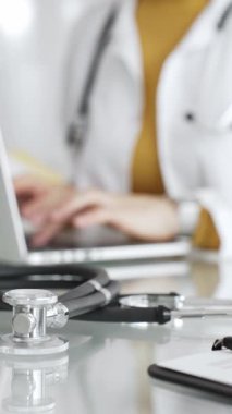 Tıbbi belgeleri olan steteskop ve pano, hasta verilerini klinikte yöneten dizüstü bilgisayar üzerinde çalışan kadın doktorun yanındaki masada. Tıp, sağlık ve bilim kavramı.