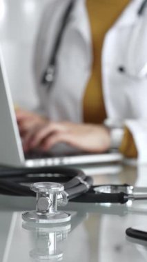 Tıbbi belgeleri olan steteskop ve pano, hasta verilerini klinikte yöneten dizüstü bilgisayar üzerinde çalışan kadın doktorun yanındaki masada. Tıp, sağlık ve bilim kavramı.