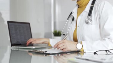 Beyaz önlüklü ve steteskoplu bir doktor bir not panosuna not yazıyor ve hasta bilgilerini kontrol ediyor. Tıp, sağlık ve bilim konsepti