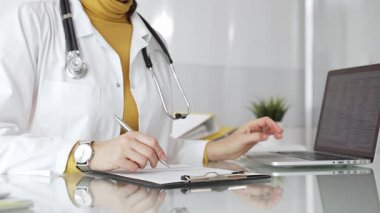 Beyaz önlüklü ve steteskoplu bir doktor bir not panosuna not yazıyor ve hasta bilgilerini kontrol ediyor. Tıp, sağlık ve bilim konsepti