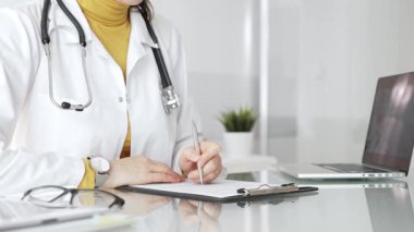 Beyaz önlüklü ve steteskoplu bir doktor bir not panosuna not yazıyor ve hasta bilgilerini kontrol ediyor. Tıp, sağlık ve bilim konsepti