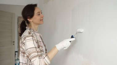 Koruyucu eldivenler giyen kadın, beyaz duvarı küçük boya rulosuyla boyuyor, iç dekorasyonu yeniliyor ve ev yenileme projesinde odayı temizliyor..