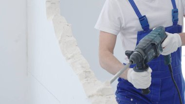 Erkek inşaat işçisi, beyaz duvarı döner çekiç matkabıyla yıkıyor, mavi tulum ve koruyucu beyaz eldivenler giyiyor, yenileme projesi sırasında toz üretiyor, yakın plan görüntüsü var..