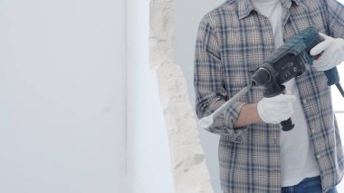 Beyaz duvarı döner çekiçle delen erkek inşaat işçisi gri kareli gömlek ve koruyucu beyaz eldiven giyiyor, yenileme projesi sırasında toz üretiyor, yakın plan