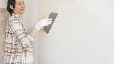 Kadın duvara macun bıçağıyla alçı yapıştırıyor, koruma için eldiven takıyor ve tadilat sırasında bile pürüzsüz ve sorunsuz bir şekilde bitmesini sağlıyor.