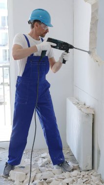 İnşaat işçisi beton duvarı döner çekiçle yıkıyor, koruyucu güvenlik giysileri giyiyor, ev yenileme çalışmaları sırasında toz ve enkaz üretiyor..