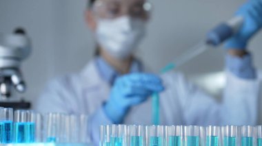 Mavi sıvıyla test tüpleri laboratuvar ortamında, yakın görüntüde. Kadın araştırmacı, arka planda bir mikro pipet kullanıyor. Tıp ve Bilim.