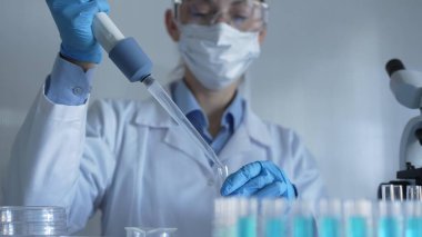 Laboratuvar önlüğü, maske, mavi eldiven ve güvenlik gözlüğü takan bilim adamı kadın test tüplerine sıvı atmak için mikropipet kullanıyor. Tıp ve Bilim.