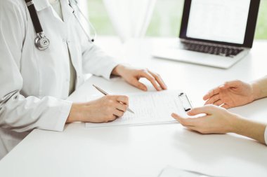 Doktor ve hasta klinikteki beyaz masada yan yana oturuyorlar. Kadın hekim bir kayıt formunu doldururken dinliyor. Tıp konsepti.