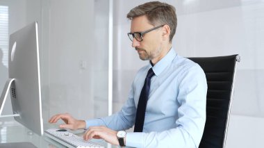 Modern bir ofisteki bir bilgisayarda çalışan işadamı, masasında yazı yazarken görevlerine odaklanıyor, profesyonelliği, verimliliği ve kurumsal bir ortamda kendini adamayı somutlaştırıyor.