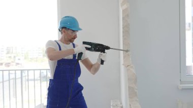Erkek inşaat işçisi beton duvarı döner çekiçle yıkıyor, iş tulumu ve mavi başlık giyiyor ev yenileme çalışmaları sırasında toz ve enkaz üretiyor, portre yatay görünümü.