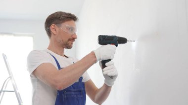 Mavi tulumlu, güvenlik gözlüklü ve iş eldivenli profesyonel inşaat işçisi kablosuz tornavida kullanarak duvara vida sokuyor..