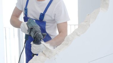 Erkek inşaat işçisi beyaz duvarı döner çekiç matkabıyla yıkıyor, mavi tulum ve koruyucu beyaz eldivenler giyiyor, yakın çekim görüntüsü var..