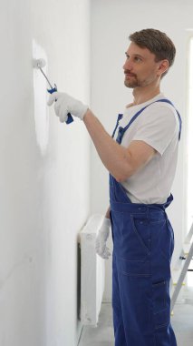 Koruyucu beyaz eldiven ve mavi iş tulumu giyen profesyonel bir ressam. Ev yenileme projesi sırasında el değmemiş iç duvarlara ruloyla boya sürüyor..