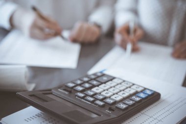 Hesap makinesi kullanılmayı beklerken, iki muhasebeci de arka planda vergi saymaya veya gelir dengesi kurmaya hazır. İş, denetim ve vergi kavramları.