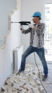İnşaat işçisi, koruyucu teçhizatlı bir çekiç matkabıyla duvarı yıkıyor ve zemin yüzeyinde inşaat enkazı oluşturuyor. Yenileme konsepti.