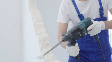 Erkek inşaat işçisi beyaz duvarı döner çekiç matkabıyla yıkıyor, mavi tulum ve koruyucu beyaz eldivenler giyiyor, yakın çekim görüntüsü var..