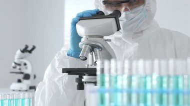 Laboratuvar ortamında araştırma yaparken mikroskop kullanarak koruyucu kıyafet giyen bir tıp bilimcisi. Test tüplerini mavi sıvıyla kapatın..