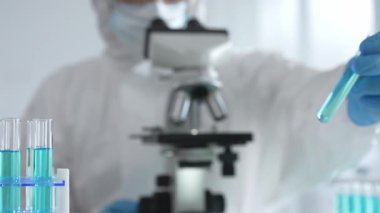 Laboratuvar ortamında araştırma yaparken mikroskop kullanarak koruyucu kıyafet giyen bir tıp bilimcisi. Test tüplerini mavi sıvıyla kapatın..
