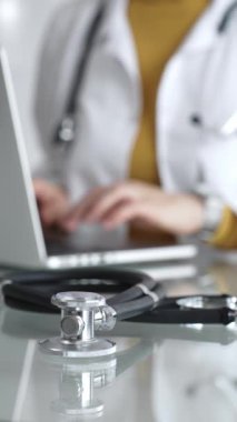 Tıbbi belgeleri olan steteskop ve pano, hasta verilerini klinikte yöneten dizüstü bilgisayar üzerinde çalışan kadın doktorun yanındaki masada. Tıp, sağlık ve bilim kavramı.