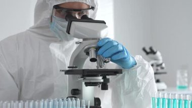 Tehlikeli madde giysisi giyen tıp virolojisti laboratuvarda mikroskopla çalışıyor. Yeni virüsler için potansiyel tedavileri araştırırken mavi sıvı örneklerini inceliyor.