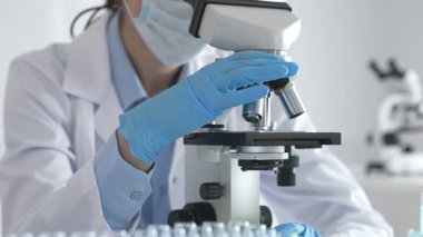 Mikroskobu ayarlayan, laboratuardaki örnekleri inceleyen, yeni ilaç veya kimyasal formülasyon araştırmaları yapan laboratuvar önlüğü giyen ve güvenlik için yüz maskesi takan bilim insanlarının eldiveni kapalı.