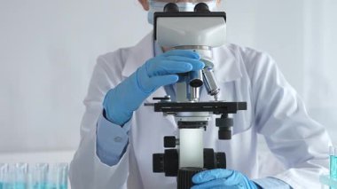 Mikroskobu ayarlayan, laboratuardaki örnekleri inceleyen, yeni ilaç veya kimyasal formülasyon araştırmaları yapan laboratuvar önlüğü giyen ve güvenlik için yüz maskesi takan bilim insanlarının eldiveni kapalı.