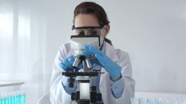 Koruyucu gözlük takan ve maske takan kadın bilim adamı mikroskop kullanarak laboratuvarda numuneleri analiz ediyor mavi sıvı test tüpleriyle çevrili yeni ilaç veya aşıları araştırıyor.