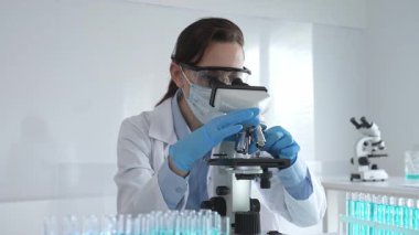 Koruyucu gözlük takan ve maske takan kadın bilim adamı mikroskop kullanarak laboratuvarda numuneleri analiz ediyor mavi sıvı test tüpleriyle çevrili yeni ilaç veya aşıları araştırıyor.
