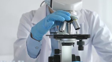 Mikroskobu ayarlayan, laboratuardaki örnekleri inceleyen, yeni ilaç veya kimyasal formülasyon araştırmaları yapan laboratuvar önlüğü giyen ve güvenlik için yüz maskesi takan bilim insanlarının eldiveni kapalı.
