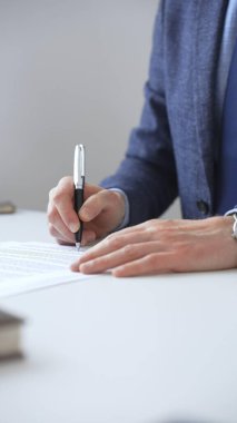 İş adamlarının zarif bir kalemle kontrat imzalaması, mavi bir takım elbise ve kol saati takması, ofisteki beyaz bir masada oturması. İş adamları konsepti.