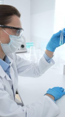 Laboratuvar önlüğü giyen ve koruyucu gözlük takan kadın bilim adamı modern laboratuarda mikroskop ve test tüpleri askısı ile test tüpünü temiz sıvıyla analiz ediyor. Tıp, sağlık ve bilim konsepti