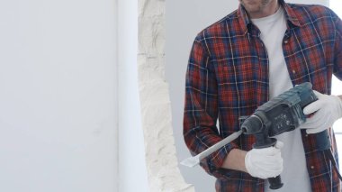 Kırmızı kareli gömlek ve koruyucu beyaz eldiven giyen erkek inşaat işçisi, yenileme projesi sırasında beyaz duvarı döner çekiçle deliyor ve toz üretiyor, yakın görüş.