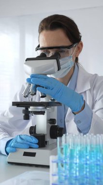 Mikroskop kaydırağını inceleyen kadın bilim adamı, laboratuarda koruyucu giysiler giyiyor, mavi renkli test tüpleriyle farmasötik buluşları araştırıyor..