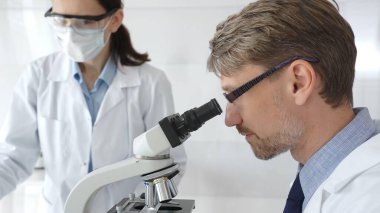 Gözlük takan bir araştırmacı mikroskop altında bir numuneyi inceliyor, koruyucu maske takan bir laboratuvar teknisyeni de çalışma sırasında beyaz laboratuvar önlükleriyle yakınlarda duruyor. Tıp ve Bilim.