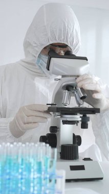 Laboratuvar ortamında araştırma yaparken mikroskop kullanarak koruyucu kıyafet giyen bir tıp bilimcisi. Test tüplerini mavi sıvıyla kapatın..