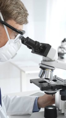 Koruyucu maskeli bir erkek bilim adamı, yeni ilaçlar ve tedaviler geliştirmek için parlak ışıklandırılmış bir laboratuvarda mikroskoba bakıyor. Tıp ve bilim konsepti.