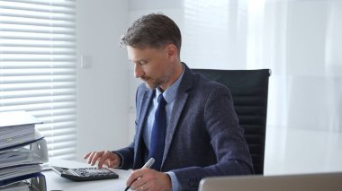 Takım elbiseli ve kravatlı olgun bir iş adamı ofis masasında oturuyor ve hesap makinesi kullanarak evrakları imzalıyor. İş dünyasında denetim ve vergi teması