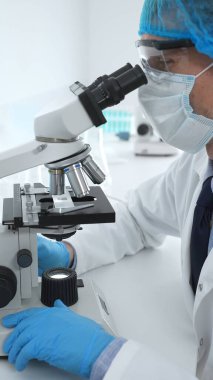 Koruyucu maske takan kıdemli erkek bilim adamı laboratuarda mikroskop kullanarak tıbbi araştırma ve analiz yapıyor. Tıp, sağlık ve bilim konsepti.